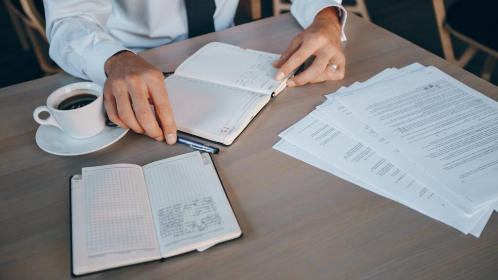 Hands rest on an open notebook while writing, with a cup of coffee and stacked documents on a wooden desk.