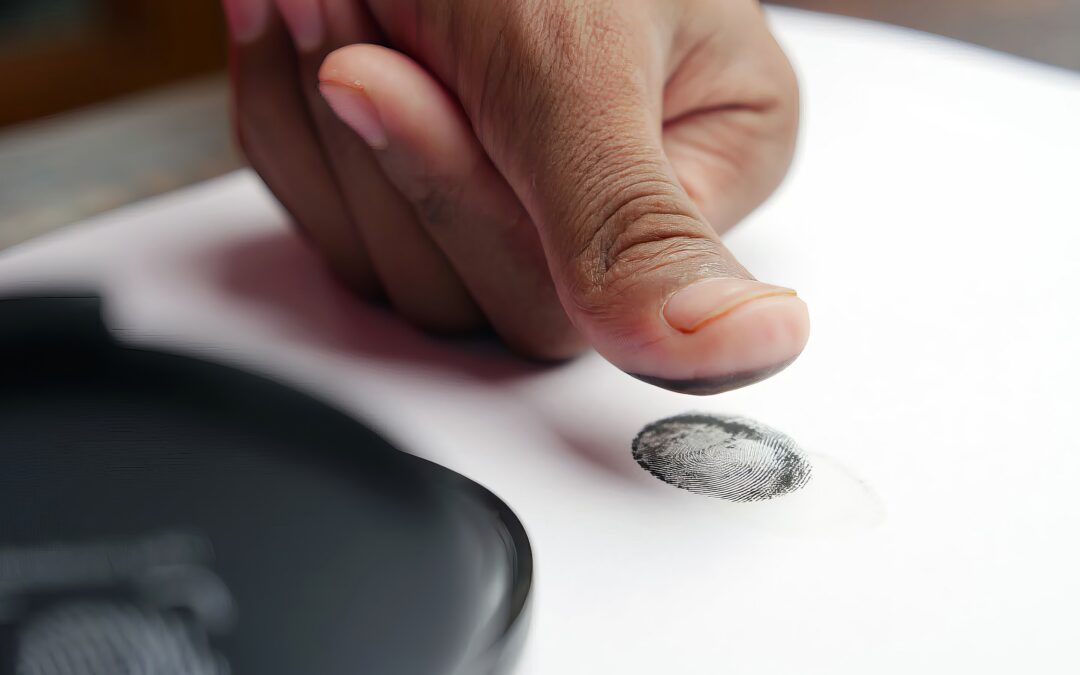 Close-up of a finger pressing a fingerprint onto a sheet of paper, with a fingerprint scanner nearby in the foreground.