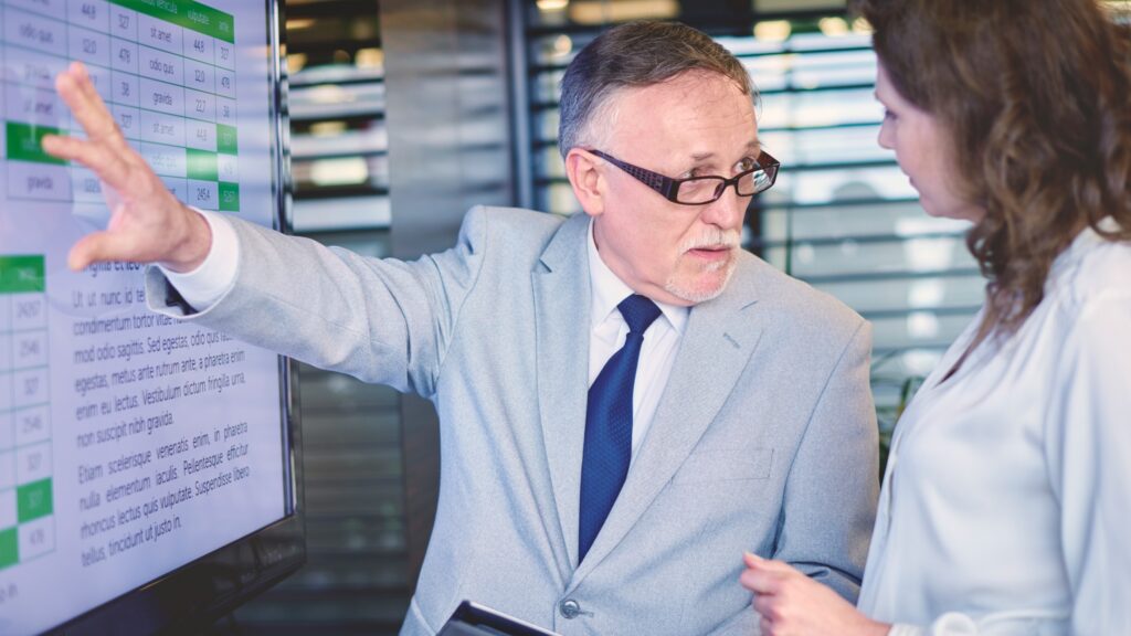Businessman in a gray suit points at a large screen showing charts and text while a colleague watches.
