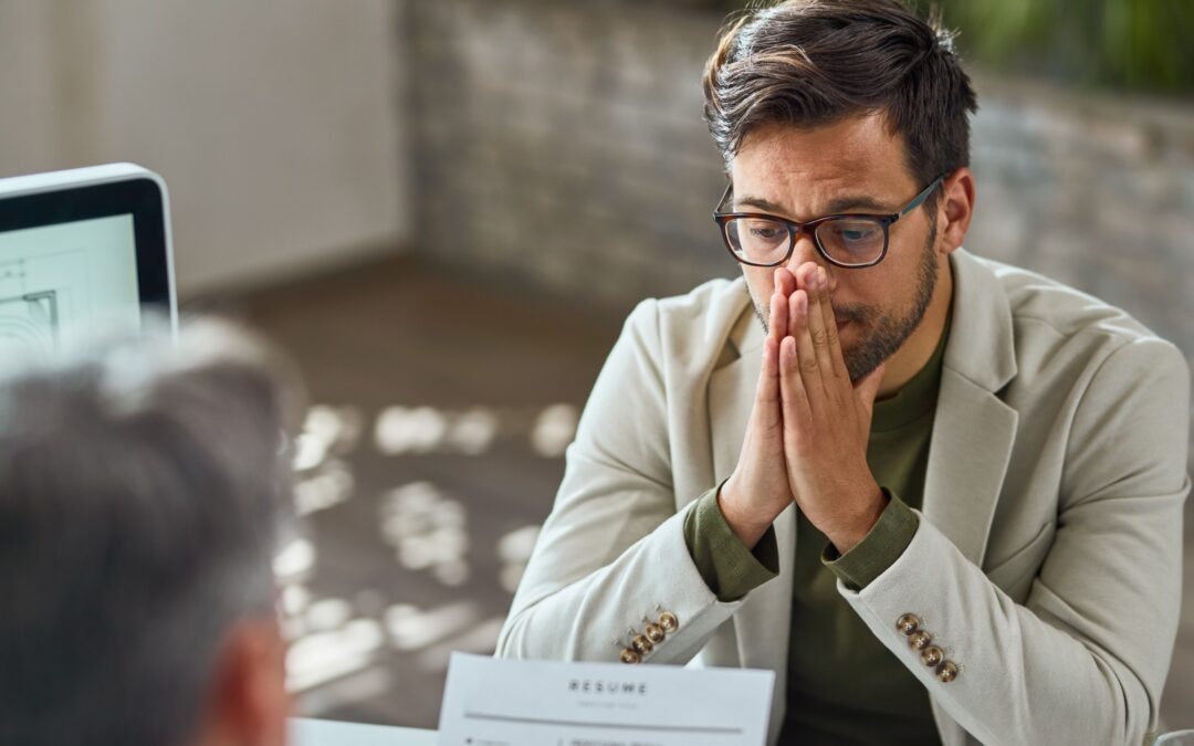 Man with glasses sits at a table, hands clasped near his mouth, appearing thoughtful during a job interview with a resume on the table.