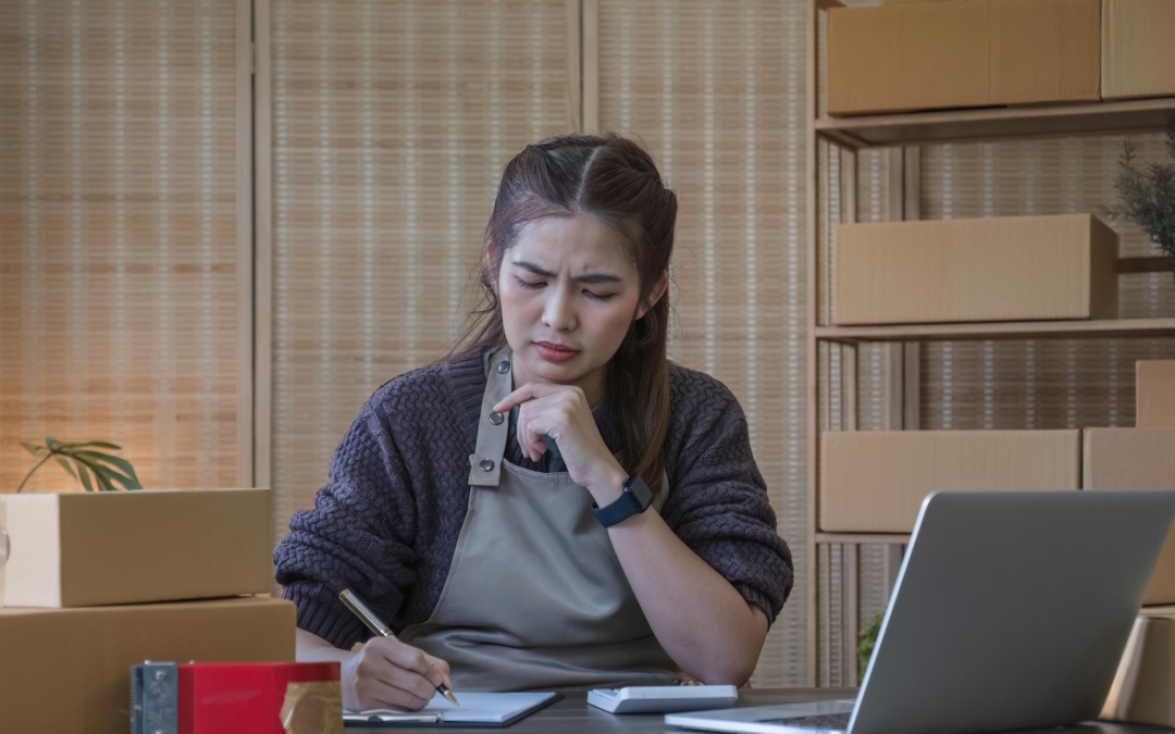 Woman in a gray apron sits at a desk, writing on a clipboard while surrounded by cardboard boxes and a laptop in a packing area.