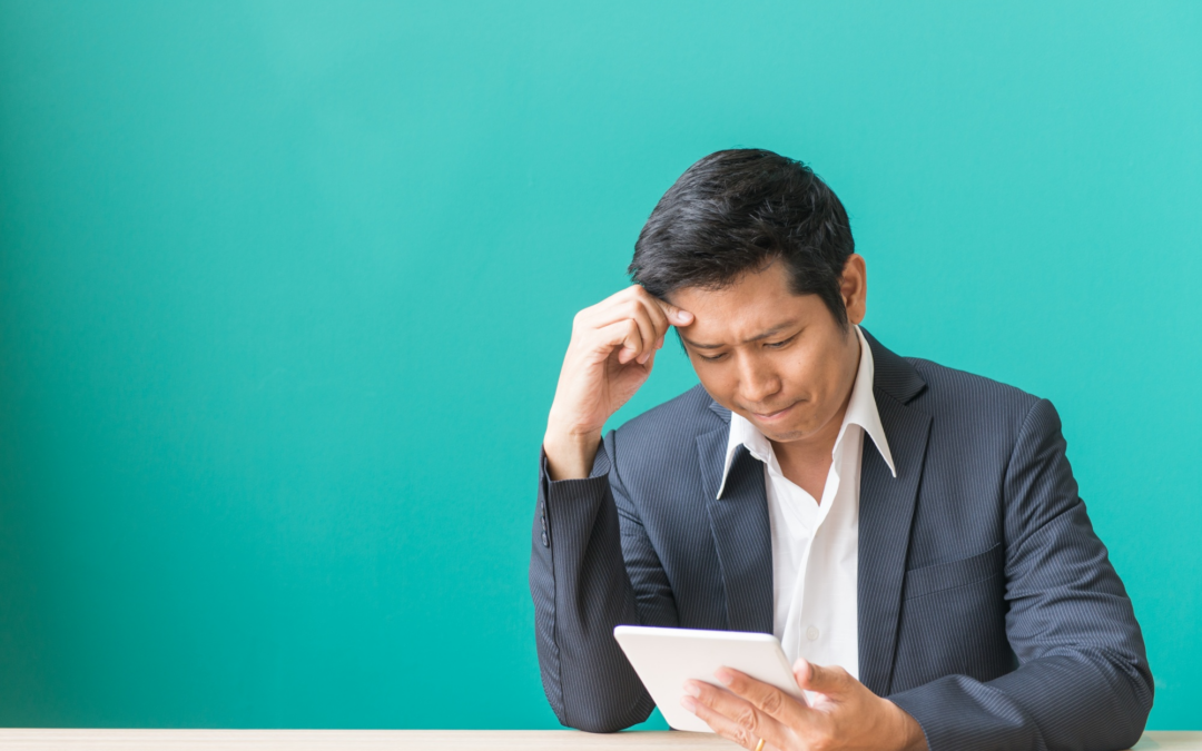 Man in a pinstripe suit sits at a desk, looking down at a tablet with a thoughtful expression against a teal background