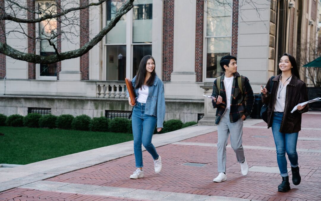 Three college students walk along a brick-paved path by a campus building, carrying notebooks and backpacks, and smiling at each other.